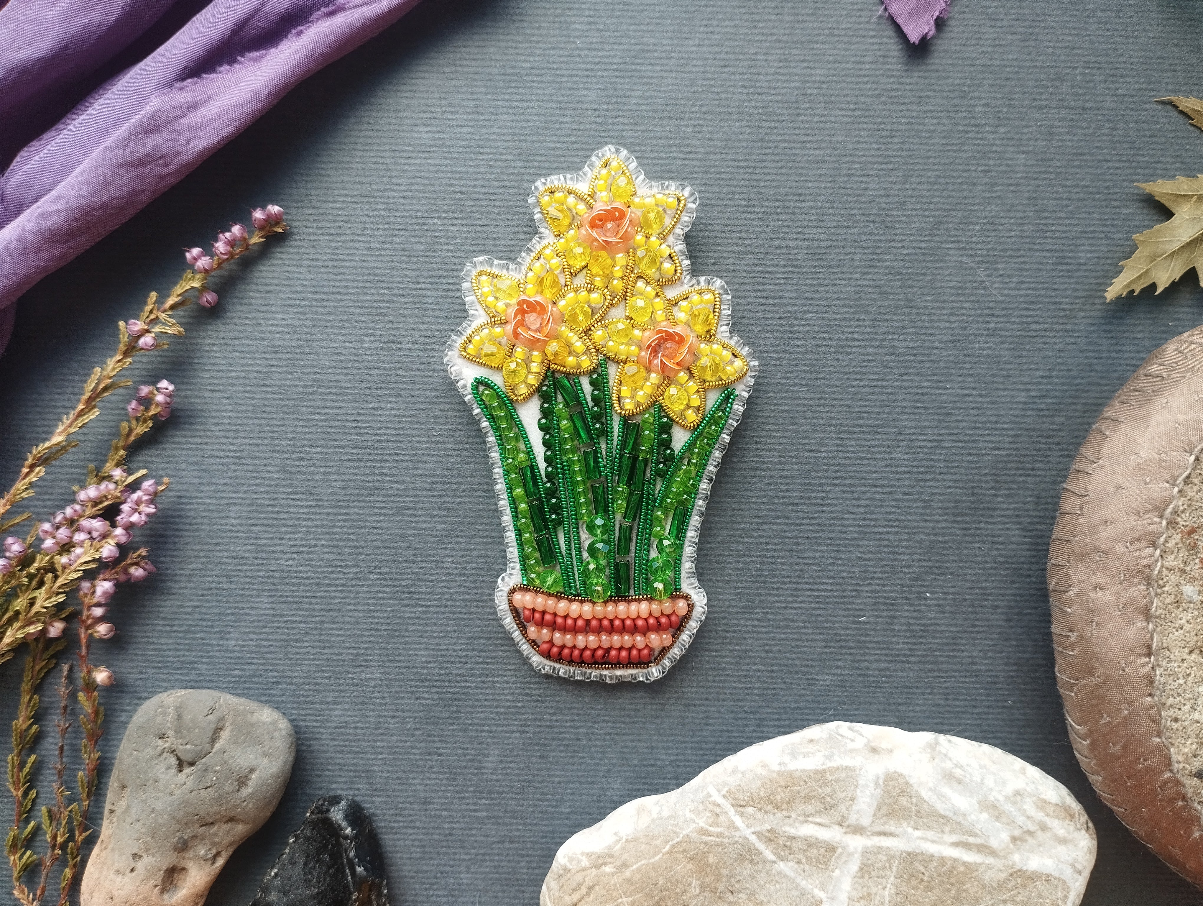 a decorative piece of embroidered flowerwork with yellow daffodils, placed on a gray background among various natural elements such as rocks, leaves, and flowers.
